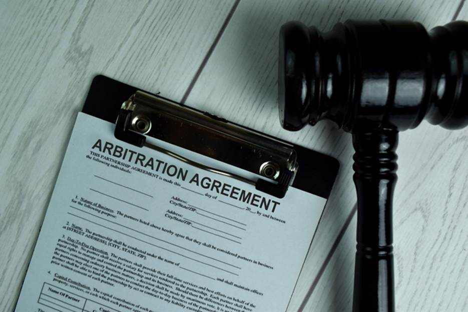 The image features an arbitration agreement form on a clipboard beside a black gavel. The form's header reads "ARBITRATION AGREEMENT."